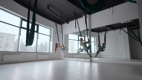 Two-young-yogi-women-doing-aerial-yoga-practice-in-green-hammocks-in-fitness-club.-Beautiful-females-working-out-in-class-performing-aero-yoga.-Variation-of-Parsvottanasana-Pyramid-pose